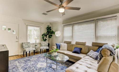 Cozy living room with a beige sectional sofa, blue accents, a glass coffee table, and natural light from large windows.