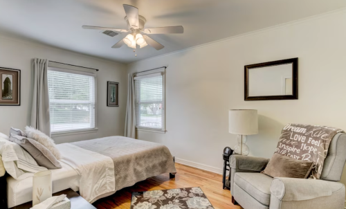Cozy bedroom with a bed, armchair, ceiling fan, and large windows, featuring warm wood floors and soft decor.