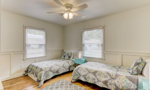 A cozy bedroom with two twin beds, a blue nightstand, and large windows with blinds.