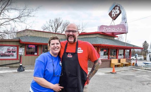 A smiling couple stands outside RB Drive-In, a retro-style diner with a red sign and outdoor seating.