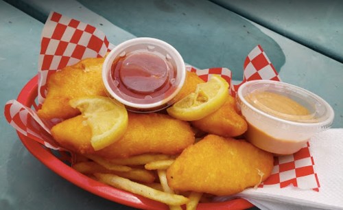 A basket of golden fried fish fillets with lemon slices, served with ketchup and a dipping sauce, alongside fries.
