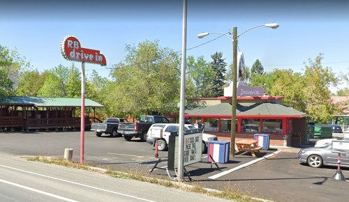 A drive-in restaurant with a red and white sign, surrounded by trees and parked cars, featuring outdoor seating.