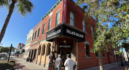 Exterior of the Palace Saloon, a historic red brick building with a black awning, surrounded by palm trees and blue sky.