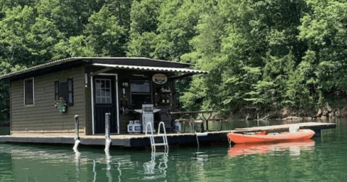 A small floating cabin on a lake, surrounded by lush green trees, with a kayak nearby.