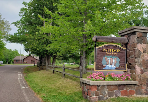 Sign for Pattison State Park with stone structure, flowers, and trees, leading to a road and park facilities in the background.