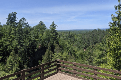 A wooden deck overlooks a lush green forest and distant hills under a clear blue sky.