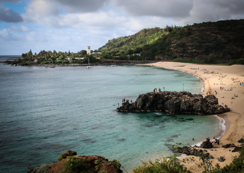 A scenic beach view with rocky outcrops, clear water, and people enjoying the sun on the sandy shore.