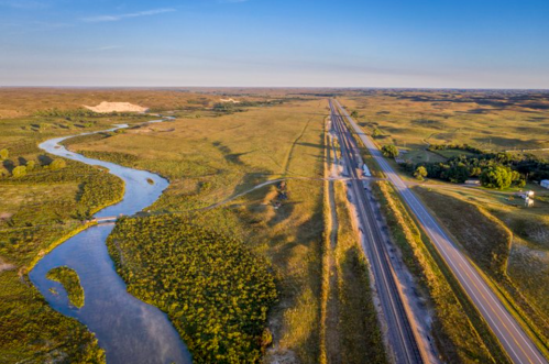 Aerial view of a winding river alongside a highway, surrounded by expansive green fields and distant hills.