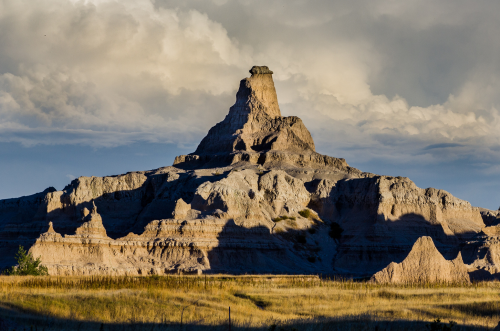 A dramatic rock formation rises against a cloudy sky, surrounded by grassy plains in a scenic landscape.