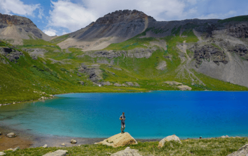 A person stands on a rock by a vibrant blue lake, surrounded by green hills and mountains under a partly cloudy sky.