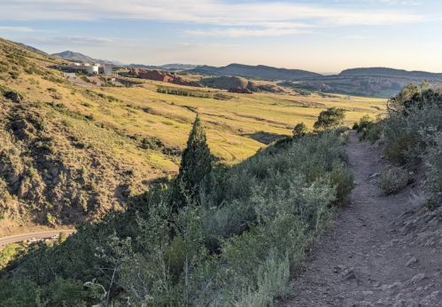 A scenic view of a winding trail overlooking a lush valley and distant mountains under a clear sky.