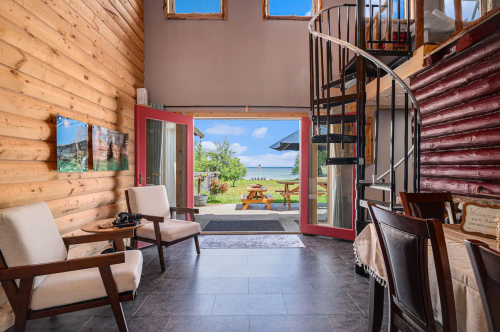 Cozy interior of a cabin with wooden walls, a spiral staircase, and a view of a patio and lake through open doors.