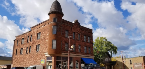 Historic brick building with a turret, under a partly cloudy sky, surrounded by trees and other structures.