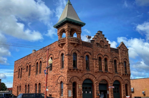 Historic red stone building with a tower, arched windows, and a blue sky with clouds in the background.