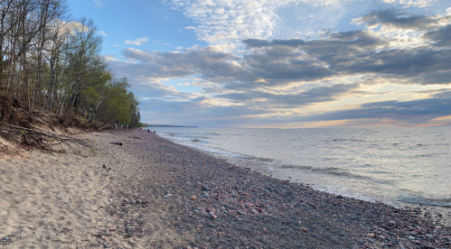 A serene beach scene with pebbles, gentle waves, and a cloudy sky, surrounded by trees along the shoreline.
