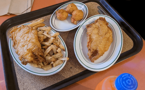 A tray with two pieces of fried fish, fries, and three hushpuppies, alongside a bottle of orange soda.