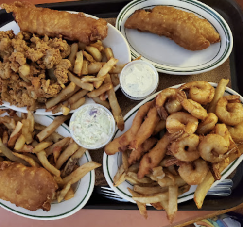 A tray of fried seafood and chicken, including shrimp, fish, fries, and coleslaw, served in white bowls.