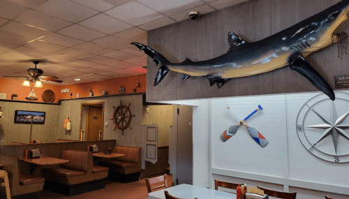 Interior of a casual restaurant featuring wooden booths, nautical decor, and a large shark mounted on the wall.