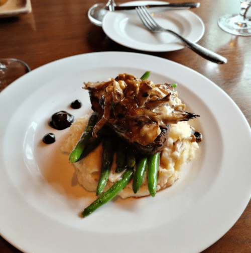 A plated dish featuring a piece of meat topped with sauce and mushrooms, served on mashed potatoes and green beans.