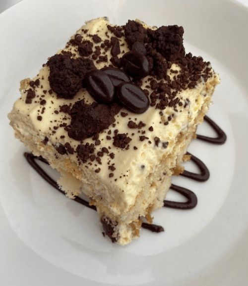 A slice of layered dessert topped with cream, chocolate crumbs, and coffee beans, drizzled with chocolate sauce.