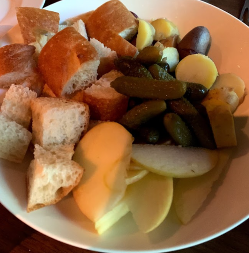 A bowl of assorted bread, pickles, olives, and sliced apples.