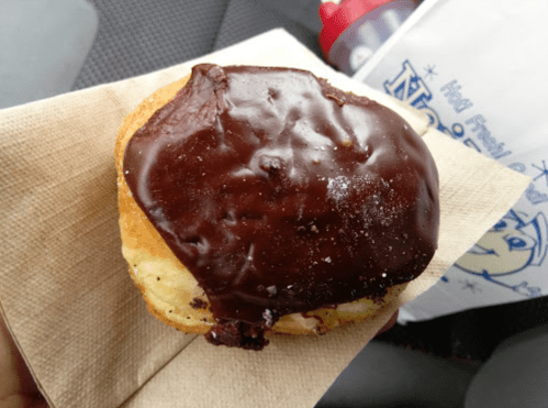 A chocolate-glazed donut resting on a napkin, held in a hand, with a bag in the background.