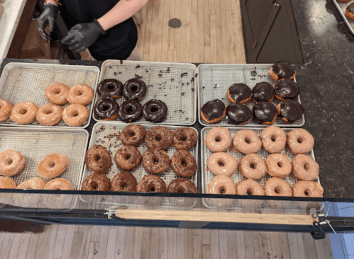 A display of assorted donuts, including glazed, chocolate-covered, and sprinkled varieties, on wire racks.