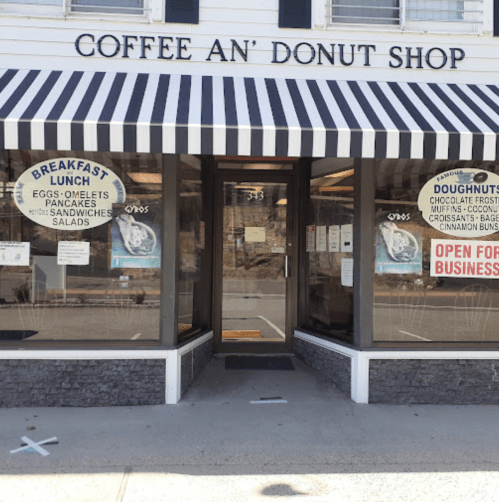 Exterior of a coffee and donut shop with a striped awning, featuring signs for breakfast and lunch options.