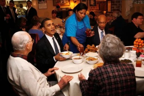 Former President Obama dining with a group of people at a restaurant, while a server brings food to the table.