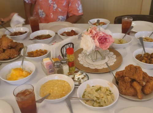 A table set with multiple bowls of Southern dishes, including fried chicken, vegetables, and drinks, with a floral centerpiece.