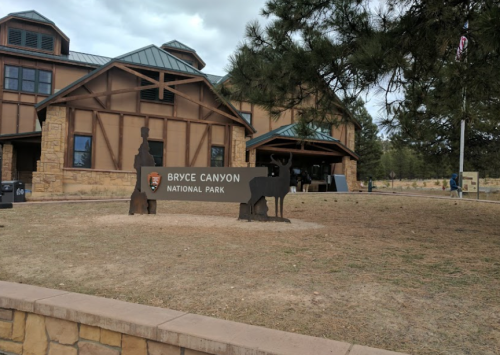 Sign for Bryce Canyon National Park in front of a visitor center with a rustic building and trees in the background.