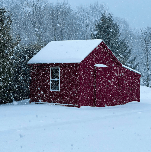 A red shed covered in snow, surrounded by a snowy landscape and falling snowflakes.
