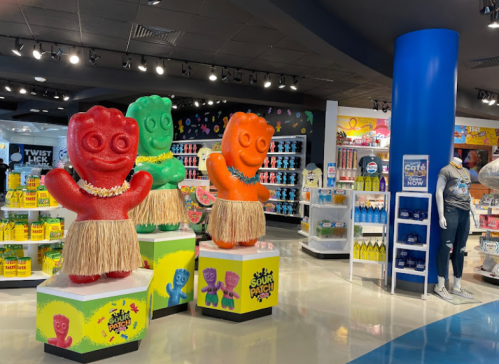 Three large gummy bear figures in hula skirts stand on pedestals in a colorful candy store. Brightly lit and vibrant decor.