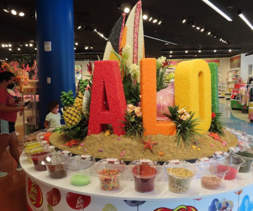 Colorful "ALO" letters surrounded by candy displays and tropical decorations in a vibrant store setting.