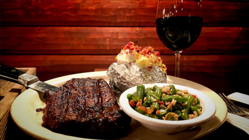 A plate with a grilled steak, baked potato topped with cheese, green beans, and a glass of red wine in the background.