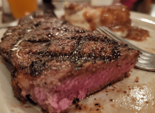 A close-up of a perfectly cooked steak with a pink center, served on a plate with a fork and a side dish in the background.
