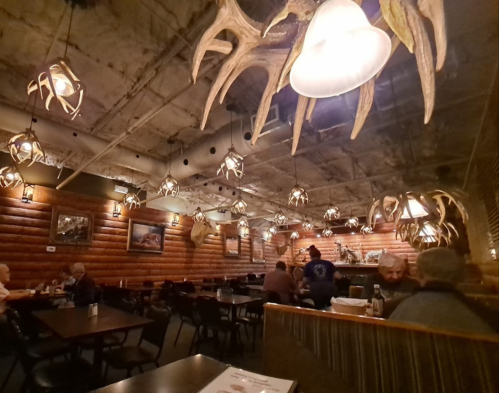 Cozy restaurant interior with wooden walls, antler light fixtures, and patrons dining at tables.