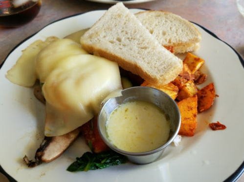 A plate of food featuring melted cheese over vegetables, roasted potatoes, and two slices of bread with a dipping sauce.