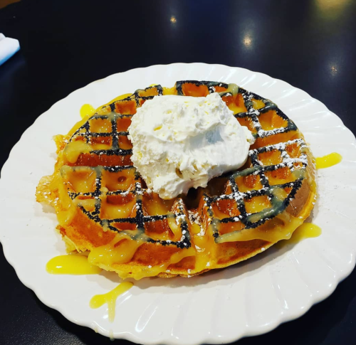 A golden waffle topped with whipped cream and drizzled with syrup on a white plate.