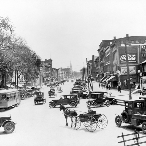 Historic street scene with early 20th-century cars and a horse-drawn carriage, bustling with activity.