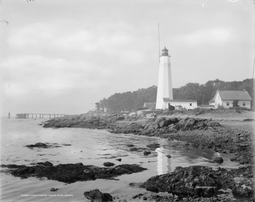 A historic lighthouse stands by the shore, with rocky terrain and a small building nearby, under a cloudy sky.