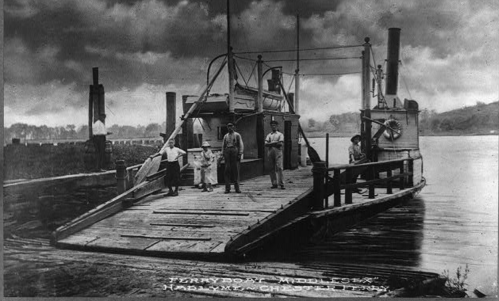 Black and white photo of a ferryboat with people standing on the dock and ramp, set against a cloudy sky.