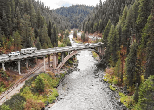 A bridge spans a river surrounded by dense forest, with vehicles traveling on the road above.