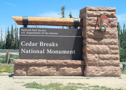 Sign for Cedar Breaks National Monument, featuring the National Park Service logo and surrounded by trees and wildflowers.