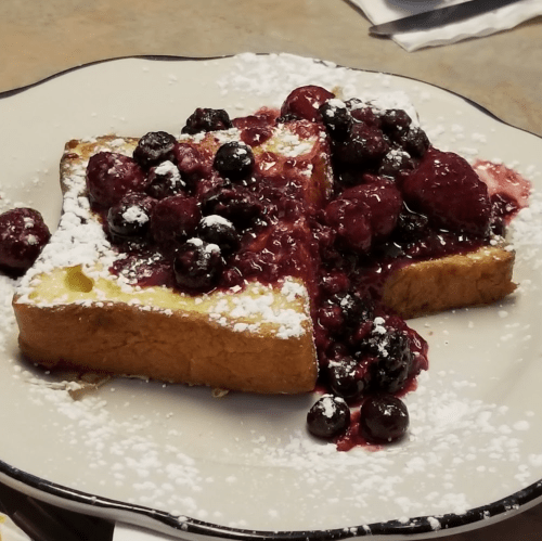 A slice of French toast topped with mixed berries and powdered sugar on a white plate.