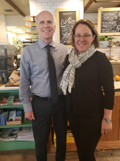 A man in a dress shirt and tie stands next to a woman in a black outfit and scarf, both smiling in a café setting.