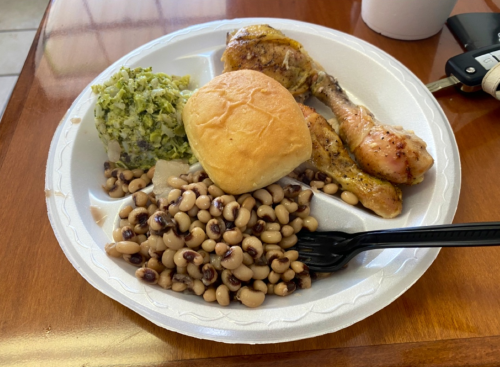 A plate of food featuring baked chicken, black-eyed peas, collard greens, and a biscuit on a white disposable plate.