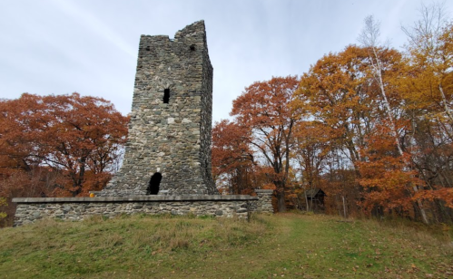 A stone tower stands amidst autumn trees with orange leaves, set against a cloudy sky.