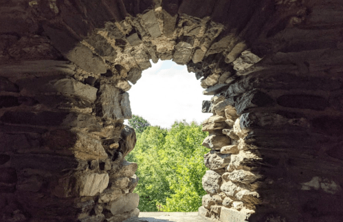 View through a stone archway, framed by rough stones, overlooking lush green trees and a bright sky.