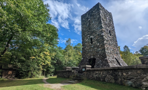 A tall stone tower surrounded by trees under a blue sky, with a path leading towards it.